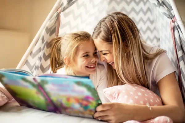 Mother and daughter giggling together while reading a kids book in a fort at home