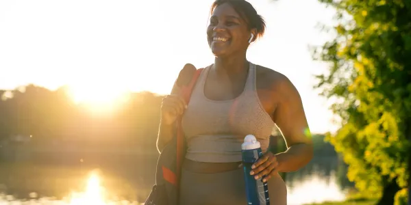 A smiling woman in workout clothes walking with a bag and water bottle in the park near a pond 